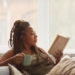Black woman seated on couch reading book while drinking something in mug - millennials homebodies